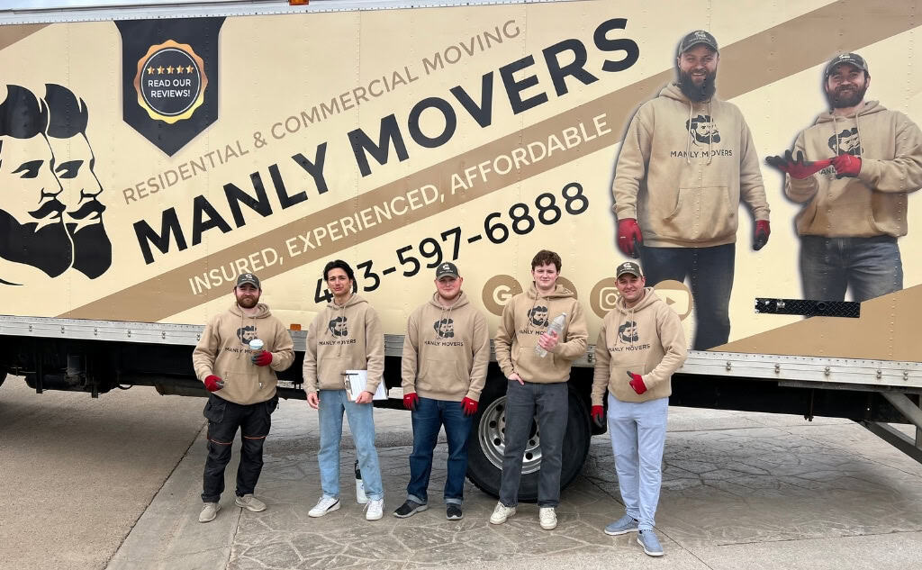 Five movers in casual attire are standing in front of a "Manly Movers" truck in Red Deer. The truck is adorned with the company's contact information and a large logo, demonstrating their expertise in packing for a move.
